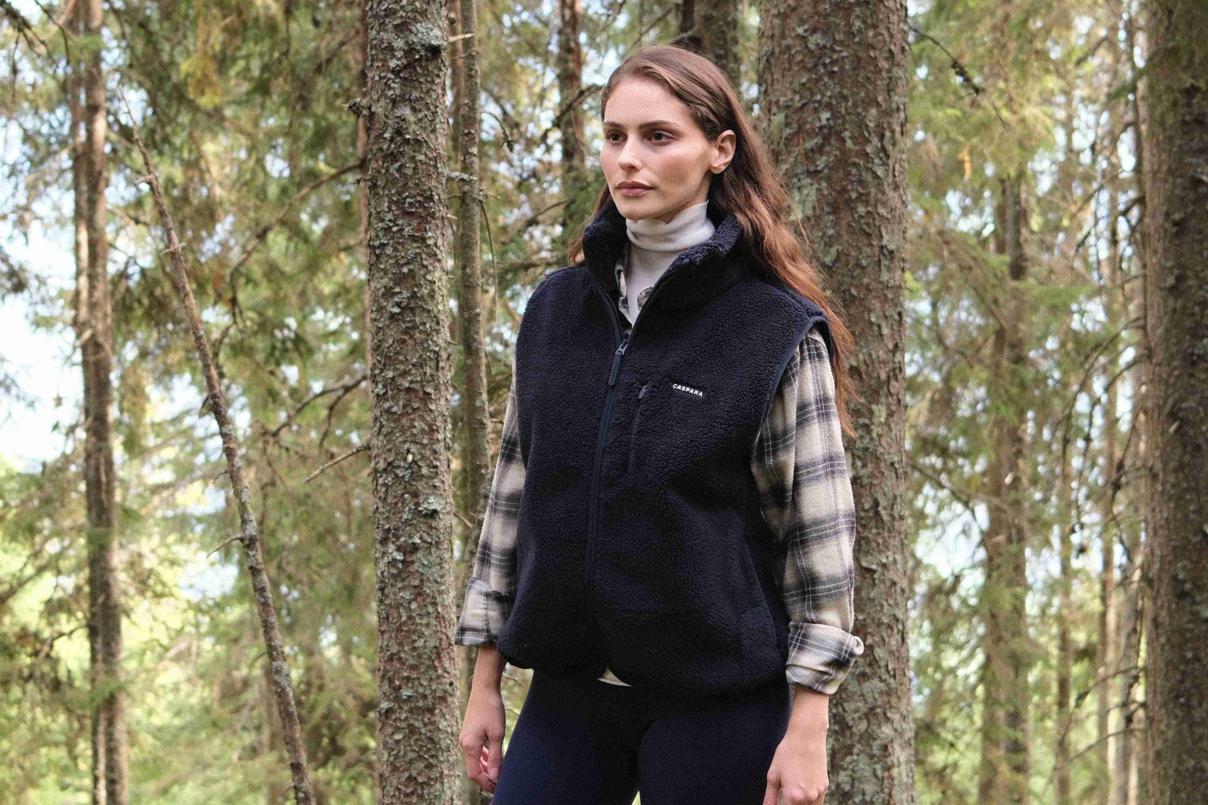 Woman wearing a navy CASPARA Cloudfleece vest outdoors in a pine forest.